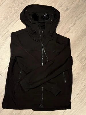Svart vindjacka från CP Company - Snygg svart vindjacka från C.P. Company med ikoniska inbyggda glasögon i huvan. Jackan har dragkedja framtill, två snedställda bröstfickor med dragkedja och justerbar huva. Tillverkad i ett slitstarkt, lätt material som passar perfekt till streetwear.