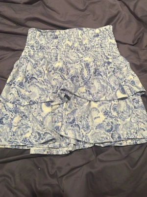 Blåvit volangkjol från Gina Tricot - Supersöt blåvit kortkjol från Gina Tricot med volangdetaljer och blommigt mönster. Kjolen har resår i midjan för skön passform och är perfekt för varma dagar. Tillverkad i mjuk bomull som känns lätt och luftig.