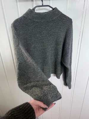 stickad tröja med hög krage - Mysig mörkgrå stickad tröja med hög krage och långa ärmar. Tröjan har en rak, något boxig passform och är tillverkad i ett mjukt, tjockt material som passar perfekt för kyliga dagar. Enkel och stilren design utan synliga loggor eller mönster.