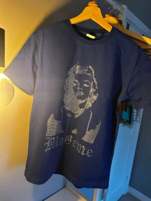 LIMITERAD mörkblå t-shirt med glittrigt tryck - Snygg mörkblå t-shirt med glittrigt tryck av Marilyn Monroe framtill. Klassisk rund halsringning och korta ärmar. Perfekt för dig som vill sticka ut med en cool och trendig look. OBS denna vara går inte längre att köpa och finns endast ett fåtal så passa på medans du kan!