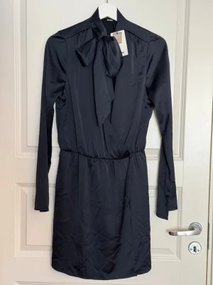 Svart klänning med knytband vid kragen - Stilren svart klänning från Bik Bok i glansigt material. Klänningen har långa ärmar och ett brett knytband i halsen som kan stylas som rosett. Resår i midjan ger en snygg passform och tyget har en elegant satinlook.