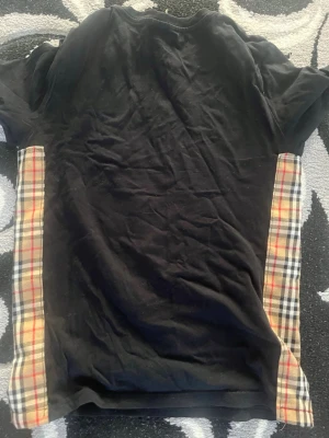 Svart t-shirt med rutiga sidopaneler - Snygg svart t-shirt med klassiska beige-rutiga sidopaneler i Burberry-inspirerat mönster. T-shirten har rund halsringning och korta ärmar. Materialet ser ut att vara bomull eller bomullsblandning. Perfekt för dig som gillar streetwear och vill sticka ut.