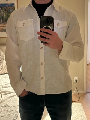 Vit Linne overshirt från Ciszere - Stilren vit skjorta från Ciszere i linne med klassisk krage, två bröstfickor med knappar och långa ärmar. Perfekt för dig som gillar en clean och avslappnad look. Skjortan har snygga träknappar och en rak passform.