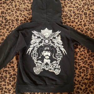 Motörhead svart hoodie med tryck - Säljer en svart hoodie från Motörhead med stort logotryck på bröstet och detaljerad vit grafik på ryggen. Tröjan har huva med snörning och en klassisk magficka. Perfekt för dig som gillar rock och vill sticka ut med en fet design.
