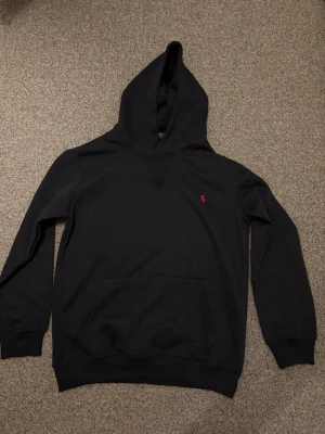 Marinblå hoodie från Polo Ralph Lauren - Marinblå hoodie från Polo Ralph Lauren med klassisk röd broderad logga på bröstet. Hoodien har huva, känguruficka och ribbade muddar vid ärmar och nederkant. Tillverkad i mjukt bomullsmaterial för en skön och avslappnad look.