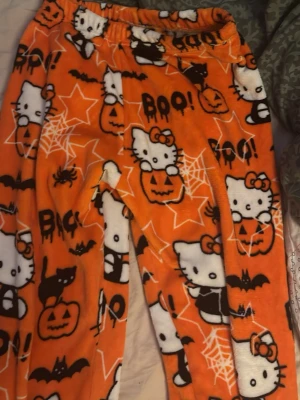 Hello Kitty Halloween-mysbyxor - Supermjuka och fluffiga orangea loungewear-byxor med Hello Kitty, pumpor, fladdermöss, spindelnät och texten 'BOO!'. Perfekta för höst och Halloween, med elastisk midja och lekfullt mönster. Materialet känns fleece-liknande och är riktigt mysigt.