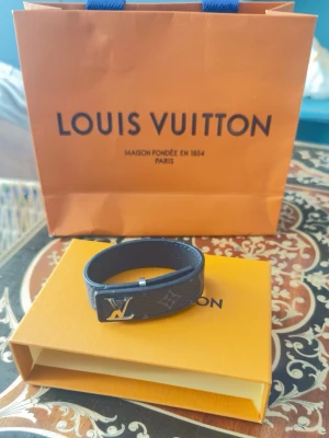 Louis Vuitton svart läderarmband - Snyggt svart armband från Louis Vuitton i präglat läder med det klassiska LV-märket i silverfärgad metall framtill. Armbandet har diskreta monogramdetaljer och en stilren design som passar perfekt till din outfit.