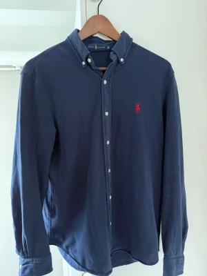 Marinblå skjorta från Ralph Lauren - Klassisk marinblå skjorta från Ralph Lauren med button-down krage och röd broderad logga på bröstet. Skjortan har långa ärmar, vita knappar och är tillverkad i mjuk bomull. Perfekt för en stilren och avslappnad look.