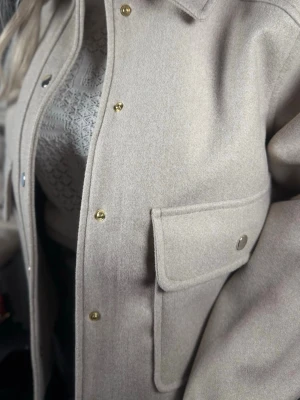 Beige overshirt med fickor M-L - Stilren beige overshirt med krage och stora bröstfickor med lock. Stängs med guldfärgade tryckknappar framtill och vid ärmslut. Overshirten har en rak passform och är tillverkad i ett mjukt, ull-liknande material.