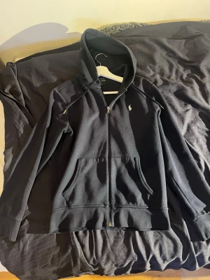 Blå hoodie från Polo Ralph Lauren - Snygg blå hoodie från Polo Ralph Lauren med dragkedja framtill och klassisk logga broderad på bröstet. Hoodien har kängurufickor, justerbar huva med snören och ribbade muddar vid ärmar och nederkant. Kan gå ner i pris 