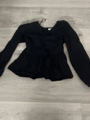 Svart topp med knytning och volang - Trendig svart topp med långa ärmar, knytning framtill och volang nedtill. 