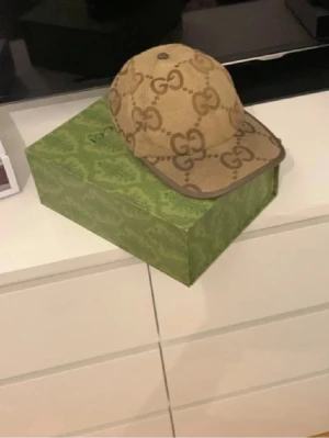 Beige GG keps från Gucci - Säljer en beige keps från Gucci med det klassiska GG-monogrammet över hela kepsen. Kepsen har bruna detaljer längs kanterna och justerbar rem bak. Materialet är canvas med insida i bomull. Perfekt för dig som vill ha en stilren och lyxig accessoar. Pris kan diskuteras✅kommer med box