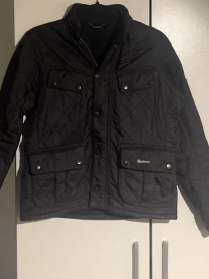 Jacka ifrån barbour - Snygg svart vindjacka från Barbour med quiltat mönster, hög krage och tryckknappar. Jackan har fyra framfickor med lock och Barbour-logga på ena fickan. Tillverkad i lätt, glansigt material som står emot vind. Perfekt för dig som vill ha en klassisk och stilren look.