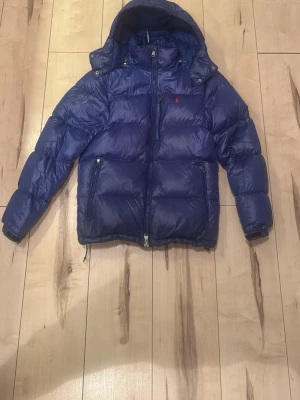 Blå pufferjacka från Polo Ralph Lauren - Blå pufferjacka från Polo Ralph Lauren med huva och dragkedja framtill. Jackan har två framfickor med dragkedja och den klassiska röda loggan broderad på bröstet. Fylld med vaddering för extra värme och har en sportig, boxig passform. Tyvärr så har den gått sönder lite på armen som man ser på bilden, har bara använt den i 1 månad, ny priss 4000kr 