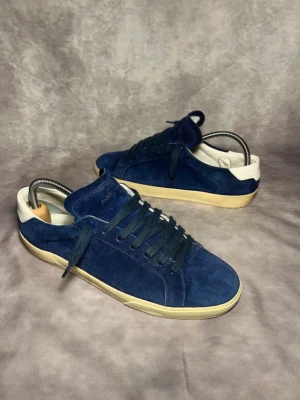 Blå mockasneakers från Saint Laurent - Snygga sneakers i blå mocka från Axel Arigato med vita detaljer vid hälen och beige sula. Klassisk låg modell med matchande blå skosnören och diskret logga på sidan. Perfekta för dig som gillar stilrena och trendiga skor.