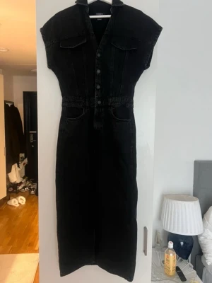 Svart jeansklänning med knappar - Snygg svart lång jeansklänning från Lindex med knappar framtill, korta ärmar och två bröstfickor. Klänningen har markerad midja, slits framtill och klassiska jeansdetaljer. Perfekt för dig som gillar en stilren och cool look.