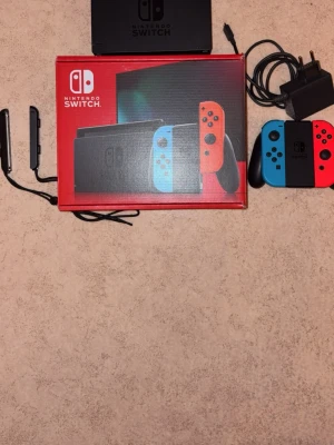 Nintendo Switch med neonröda och blå Joy-Con - Nintendo Switch spelkonsol med neonröda och blå Joy-Con-kontroller. Konsolen har en svart rektangulär form och levereras med originalkartong, laddare, dockningsstation och handkontrollhållare. Nästan aldrig använd och kan helt enkelt säga att den är helt ny.