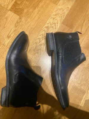 Svarta Chelsea boots från Bugatti - Snygga svarta Chelsea boots från Bugatti i slätt läder med elastiska sidopaneler och låg klack. Klassisk siluett med dragflik bak för enkel påtagning. Perfekta för en stilren och modern look.