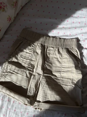 Beiga shorts med resårmidja - beiga shorts med bred resår i midjan och uppvikta benslut. Shortsen har två sidofickor och är gjorda i ett lätt, vävt material som känns luftigt. Perfekta för varma dagar och enkel att matcha med olika stilar.