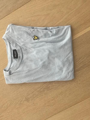 Ljusgrå t-shirt från Lyle & Scott - Snygg ljusgrå t-shirt från Lyle & Scott med klassisk passform och gul logga på bröstet. Tillverkad i mjuk bomull och perfekt för en clean och stilren look.