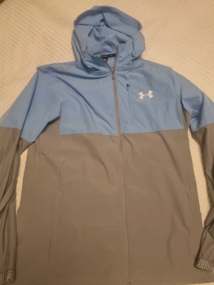 Under Armour vindjacka blå/grå - Snygg vindjacka från Under Armour i ljusblått och grått. Jackan har huva, hel dragkedja och en liten bröstficka med dragkedja. Perfekt för blåsiga dagar och sportiga outfits. Under Armour-logga på bröstet.