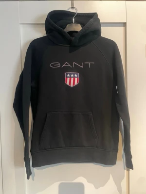Svart hoodie från GANT med logga - Svart hoodie från GANT med broderad logga och sköld på bröstet. Klassisk känguruficka framtill och stor huva. Mjuk bomullsblandning och avslappnad passform, perfekt för en chill look.