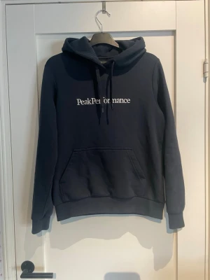 Svart hoodie Peak Performance - Svart hoodie från Peak Performance med vit logga på bröstet. Klassisk modell med huva och dragsnören samt en stor magficka framtill. Mjuk och skön bomullsblandning, perfekt för en avslappnad stil. Den ända defektern är på ena snöret så har den tappat en svart sak som ska sitta på snöret. Skriv om ni vill se närmare bild 