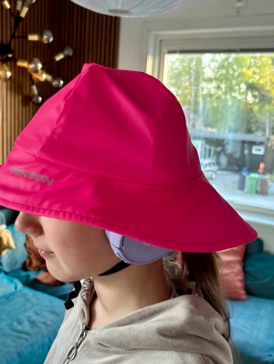 Rosa  regnhatt med bred brätte - Säljer en  regnhatt med bred brätte, perfekt för att hålla dig torr under regniga dagar. Hatten är tillverkad i ett vattenavvisande material och har en klassisk bucket hat-form. Snygg och praktisk accessoar för dig som vill sticka ut.