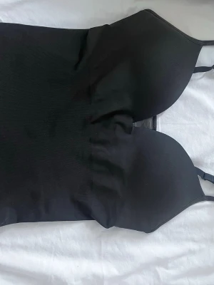 Svart body med tunna axelband - Snygg svart body med vadderade kupor och justerbara tunna axelband. Kroppsnära passform och slät yta, perfekt att bära under kläder eller som ett statement-plagg. Materialet känns stretchigt och bekvämt.