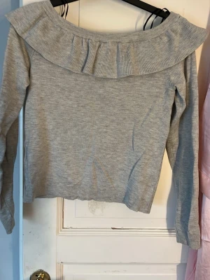 Ljusgrå stickad tröja med volang - Supersöt ljusgrå stickad tröja med bred volang runt axlarna. Tröjan har lång ärm och en mjuk, följsam passform. Perfekt för dig som gillar en feminin och trendig look. Materialet känns mjukt och bekvämt mot huden.