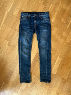 Mörkblå Lean Dean jeans från Nudie - Snygga mörkblå Lean Dean jeans från Nudie med klassiska orange sömmar och fem fickor. Jeansen har en lätt tvättad look med slitningar framtill och Nudie-logga på bakfickan. Perfekta för en avslappnad och trendig stil. Storlek 31/34.