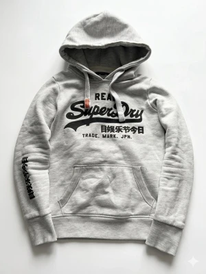 Grå hoodie från Superdry - Snygg grå hoodie från Superdry med svart tryck på bröstet och japanska tecken på ärmen. Klassisk känguruficka och dragsko i huvan. Mjuk insida och ribbade muddar. Perfekt för en avslappnad streetstyle-look. Skick 9/10 finns litet märke på armen, storlek xss/ xs, priset inte huggt i sten.