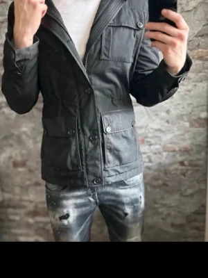 Svart/grå field jacket - Snygg svart grå field jacket med fyra stora fickor framtill och knappstängning. Jackan har en klassisk krage och är långärmad, perfekt för dig som vill ha en stilren och funktionell look. Passar till många olika outfits och har en modern, rak passform.