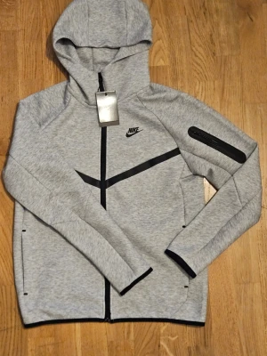 Grå Nike Tech Fleece hoodie - Snygg grå Nike Tech Fleece hoodie med svart dragkedja och svarta detaljer. Hoodien har huva, fickor och en cool zip-ficka på ärmen. Tillverkad i mjukt och stretchigt material som passar perfekt till streetwear-stilen. Nike-logga på bröstet.
