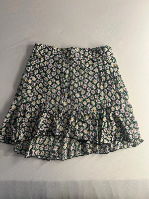 Blommig volangkjol med dragkedja - Kort kjol med blommigt mönster i vitt, gult och lila på svart botten. Kjolen har volangdetalj nedtill och dragkedja i sidan. Perfekt för dig som gillar färgglada och lekfulla plagg.