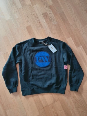Svart sweatshirt från Pernilla Wahlgren - Cool svart sweatshirt från Pernilla Wahlgren med blått PW-logo broderat på bröstet. Tröjan har rund halsringning, ribbade muddar och är tillverkad i mjuk bomull. Perfekt för en avslappnad och trendig stil.
