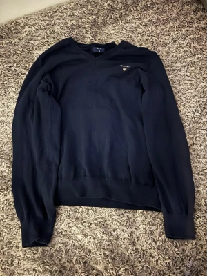 Mörkblå V-ringad tröja från GANT - Klassisk mörkblå V-ringad tröja från GANT med broderad logga och sköld på bröstet. Tröjan har långa ärmar, ribbade muddar och är tillverkad i mjukt material som passar perfekt till jeans eller skjorta.