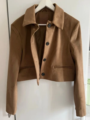 Brun mocka jacka  - Snygg brunocka imitation jacka med klassisk krage och tre knappar framtill. Jackan är kortare i modellen och har raka fickor utan lock. Perfekt för dig som vill ha en stilren och trendig look med en tidlös touch. Jag är vanligtvis en 36/38 men tog denna i 40 då den är väldigt liten i storlek! Aldrig använd. 