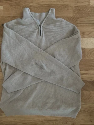Beige stickad halvzip tröja - Snygg beige stickad tröja med halv dragkedja och hög krage. Tröjan har långa ärmar och ribbstickade muddar vid ärmslut och nederkant. Perfekt för lager-på-lager och enkel att matcha med andra plagg.