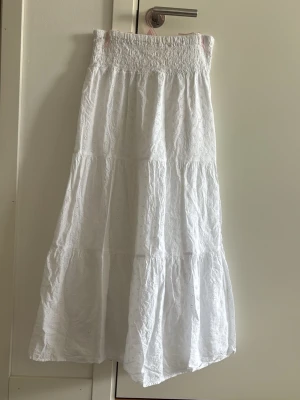 Vit lång kjol från Gina Tricot - Säljer en vit lång kjol från Gina Tricot med smock i midjan och broderade detaljer. Kjolen har en luftig och flowy passform med flera volanger, perfekt för en somrig och avslappnad stil.