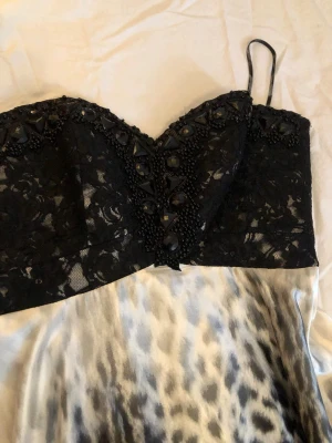 Svart spetsklänning med leopardmönster - Snygg lång klänning med svart spetsliv och dekorativa stenar upptill. Kjolen har ett iögonfallande leopardmönster i svart, grått och vitt med  snörning bak. Klänningen är  perfekt för dig som vill sticka ut och samtidigt ha en design som är ger dig en både elegant och unik look.