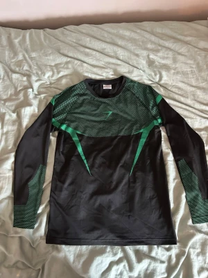 Svart och grön långärmad träningströja - Snygg långärmad träningströja från Gymshark i svart med gröna detaljer och mönster. Tröjan har rund halsringning och är tillverkad i ett stretchigt funktionsmaterial som andas, perfekt för gym eller löpning.