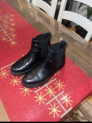 Svarta Chelsea boots i skinn - Säljer ett par klassiska svarta Chelsea boots i slätt skinn med elastiska paneler på sidorna. Bootsens design är stilren och tidlös med rundad tå och låg klack. Perfekta för dig som vill ha en enkel men snygg look till hösten eller vintern.