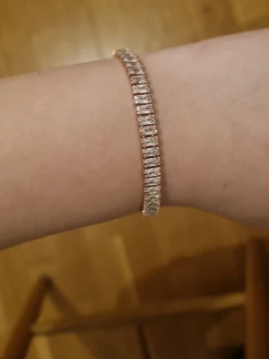 Guldigt armband med glittrande stenar - Snyggt armband i guldig metall med rektangulära, glittrande stenar längs hela bandet. Armbandet har en klassisk och stilren design med smidig låsning. Perfekt för dig som gillar att sticka ut med lite extra bling.