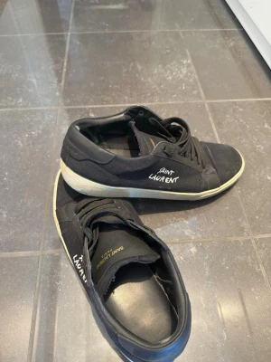 Svarta Saint Laurent sneakers - Svarta sneakers från Saint Laurent med vit sula och handskriven logga på sidan. Skorna har klassisk låg modell, svarta skosnören och ovandel i canvas och mocka. Perfekta för en stilren och trendig look. Storlek 45. Box och dustbag ingår självklart!