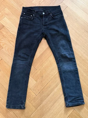 Svarta jeans från Nudie - Snygga svarta jeans från Nudie med klassisk femficksdesign och subtila slitningar. Modell: Grim Tim. Storlek: 28/28.  Jeansen har normal midja och raka ben, tillverkade i mjuk bomullsdenim med stretch för extra komfort.