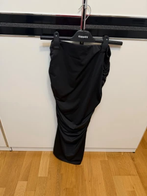 Svart långkjol med rynkade detaljer - Snygg svart långkjol med rynkade detaljer längs ena sidan som ger en cool och trendig look. Kjolen har en smal passform och är tillverkad i ett mjukt och stretchigt material som sitter bekvämt. 