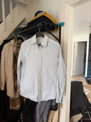 Ljusblå randig skjorta These Glory Days - Snygg ljusblå skjorta från These Glory Days med tunna vita ränder. Skjortan har klassisk krage, knäppning framtill och lång ärm. Slim fit-modell i mjuk bomull, perfekt för en clean och stilren look.