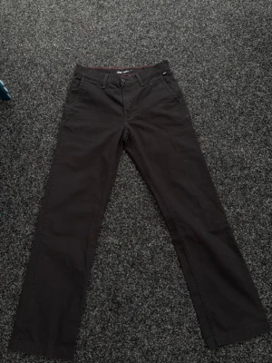 Svarta chinos från Vans - Snygga svarta chinos från Vans med relaxed fit och röda sömmar på insidan av linningen. Klassisk femficksmodell med knapp och dragkedja. Perfekta för en avslappnad och stilren look.