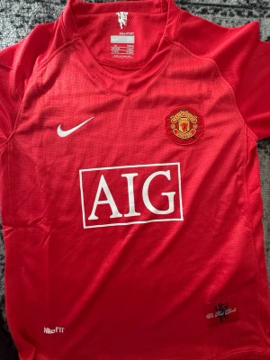 Manchester United matchtröja Nike - Röd Manchester United matchtröja från Nike med klubbmärke och AIG-logga på bröstet. Tröjan har korta ärmar, V-ringning och är tillverkad i lätt, ventilerande material. Perfekt för fotbollsträning eller att bära på läktaren.